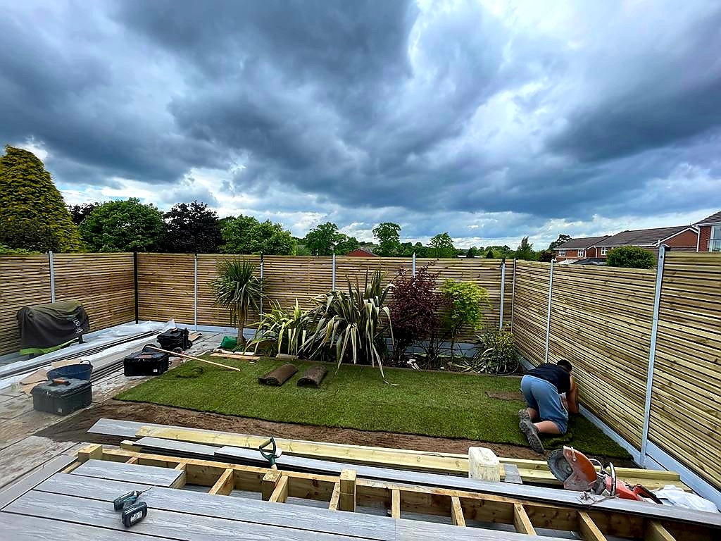 A rear garden mid-transformation — artificial lawn being laid over a prepared base, new timber fencing up and decking joists set out, under a moody Yorkshire sky.