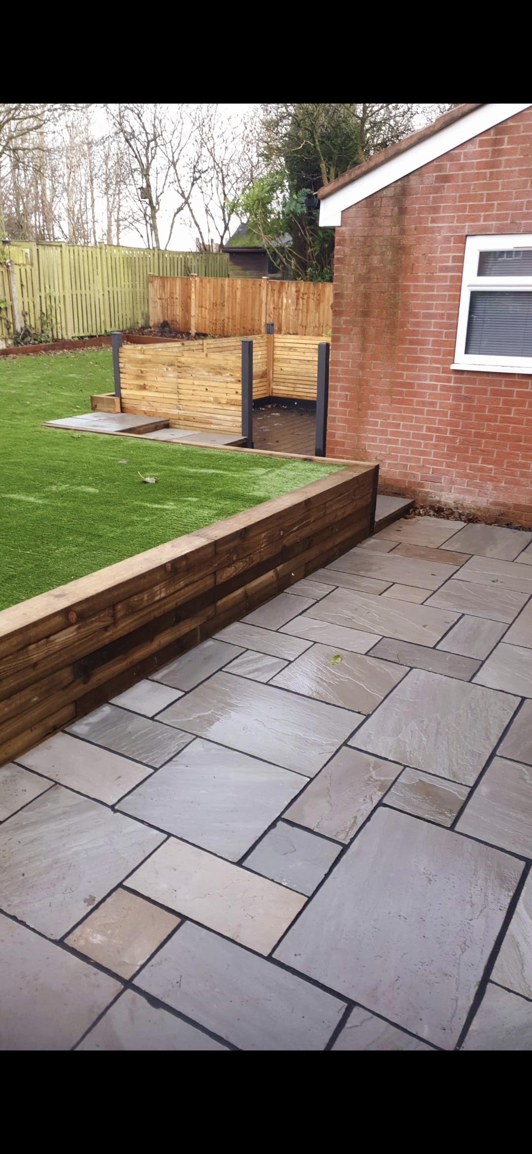 A grey Indian-sandstone patio laid to falls, framed by a timber-sleeper retaining wall that holds a raised artificial lawn — hard and soft landscaping integrated in one scheme.